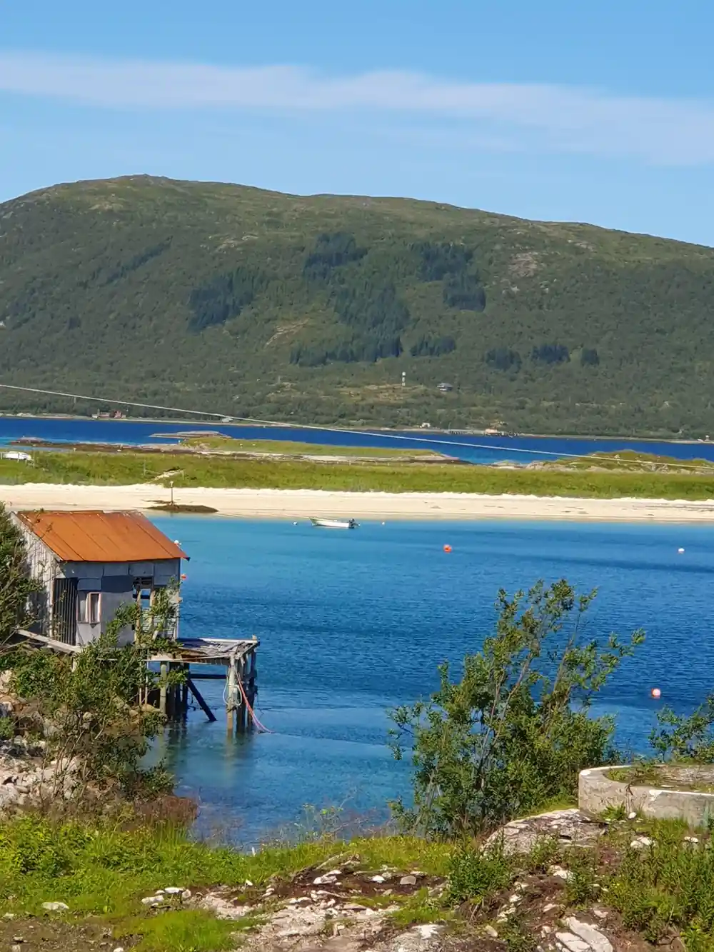 A boathouse on stilts beside a white beach and green hill
